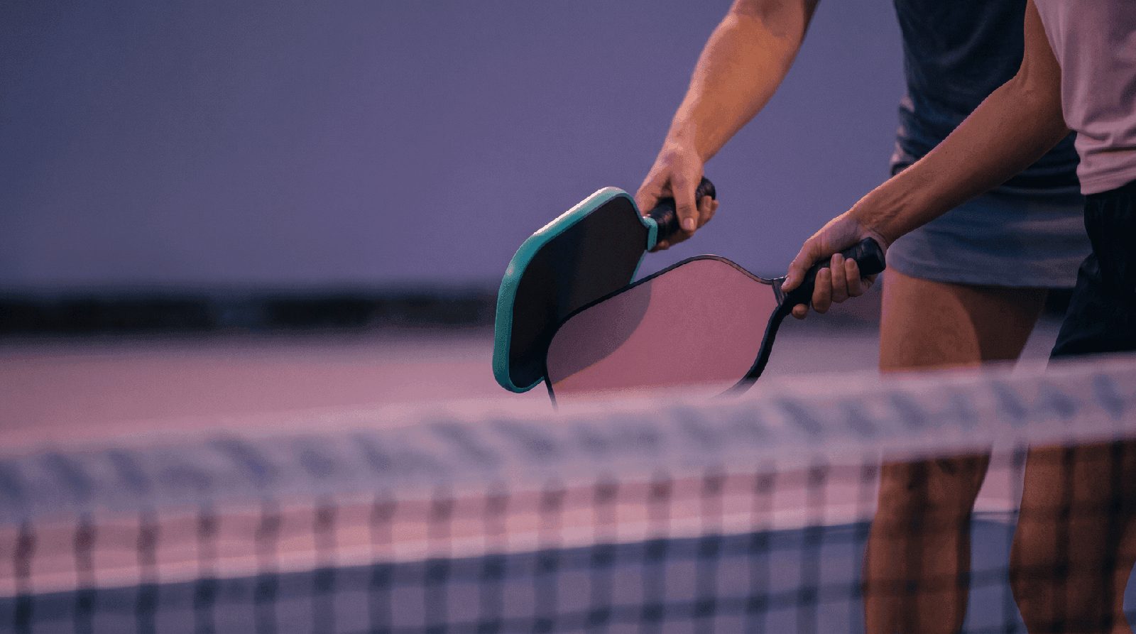 Close-up of two pickleball paddles tapped together at the net in the "good game" gesture, dusk light, no faces visible.